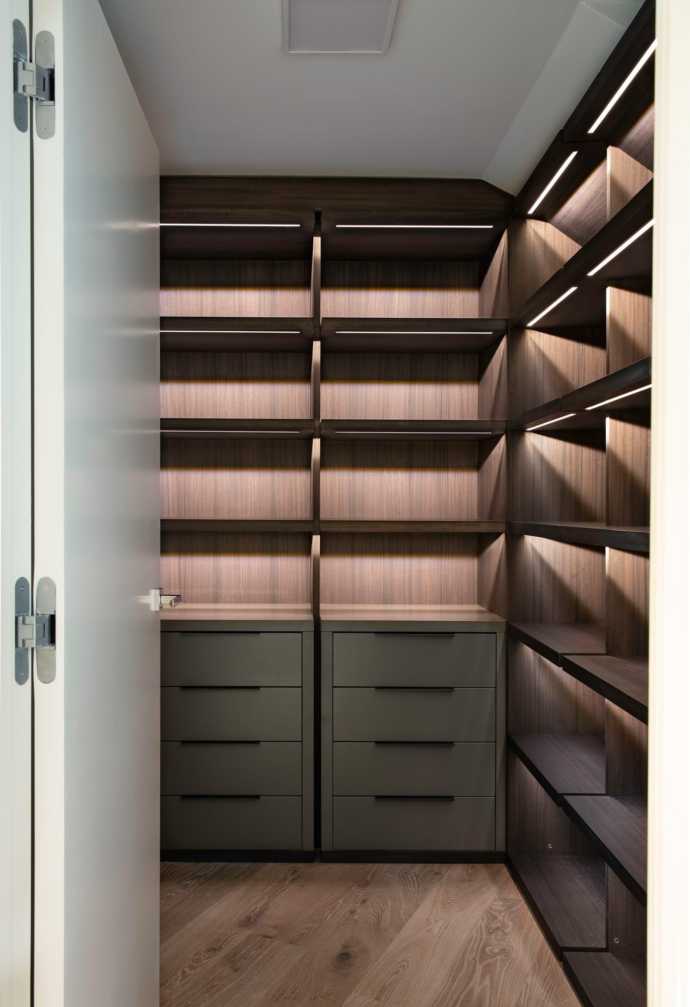 Modern dark wood walk-in closet with integrated LED shelf lighting and grey drawers. Modern dark wood walk-in closet with integrated LED shelf lighting and grey drawers.