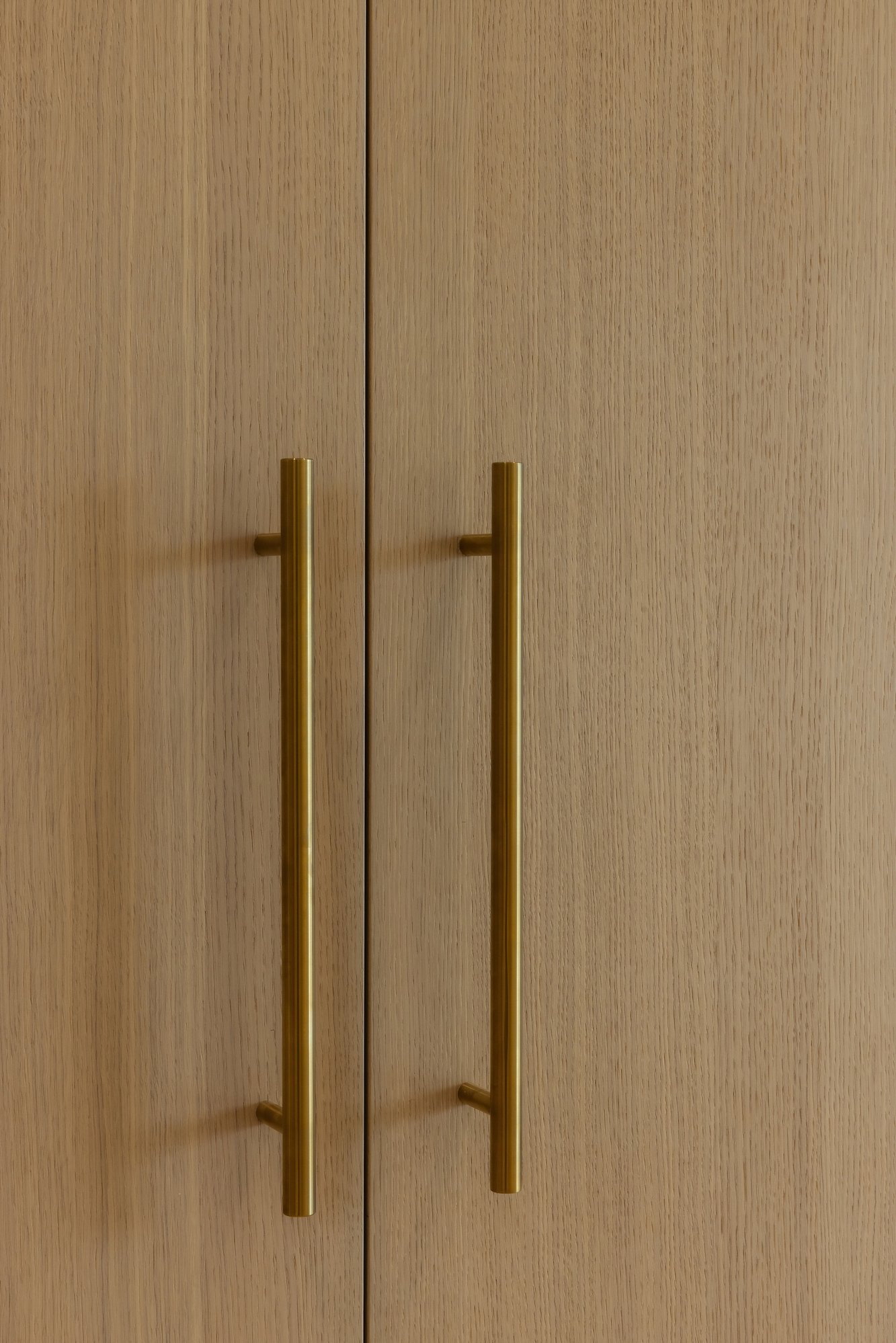 Close-up of light oak wood cabinetry with vertical brushed gold bar handles. Close-up of light oak wood cabinetry with vertical brushed gold bar handles.