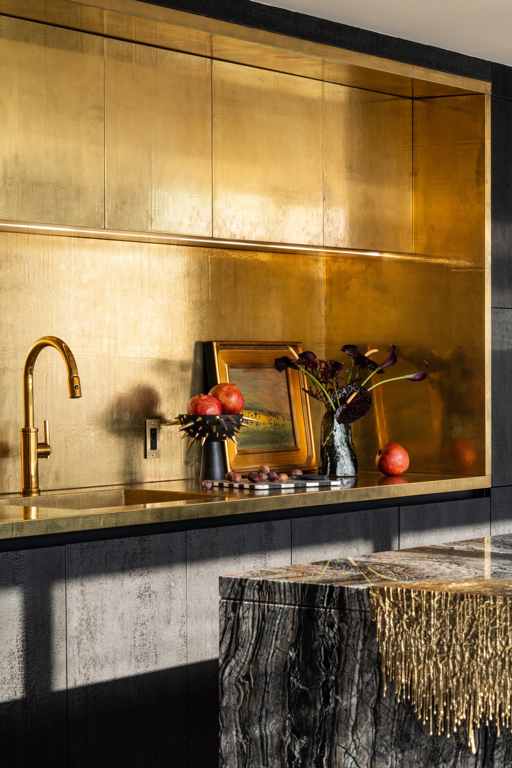 Luxury kitchen featuring a textured gold metal backsplash and recessed upper cabinetry, a gold faucet and sink, and a dark marble island with unique gold fringe detailing. Luxury kitchen featuring a textured gold metal backsplash and recessed upper cabinetry, a gold faucet and sink, and a dark marble island with unique gold fringe detailing.