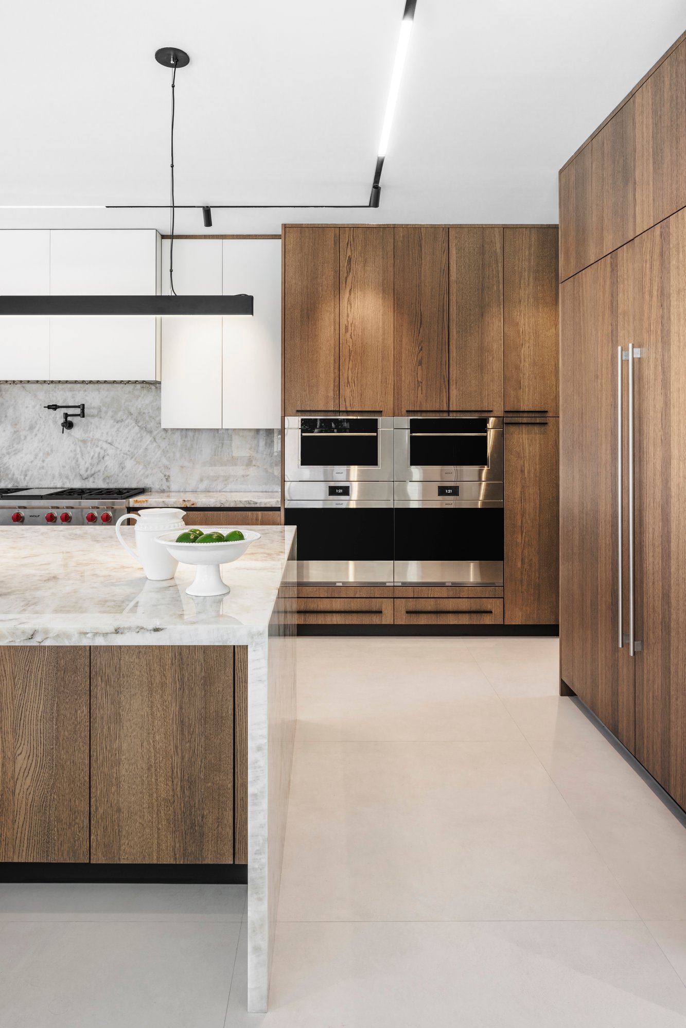 Luxury kitchen featuring a white marble waterfall island, natural wood grain cabinetry, and integrated Wolf wall ovens with a marble backsplash. Luxury kitchen featuring a white marble waterfall island, natural wood grain cabinetry, and integrated Wolf wall ovens with a marble backsplash.