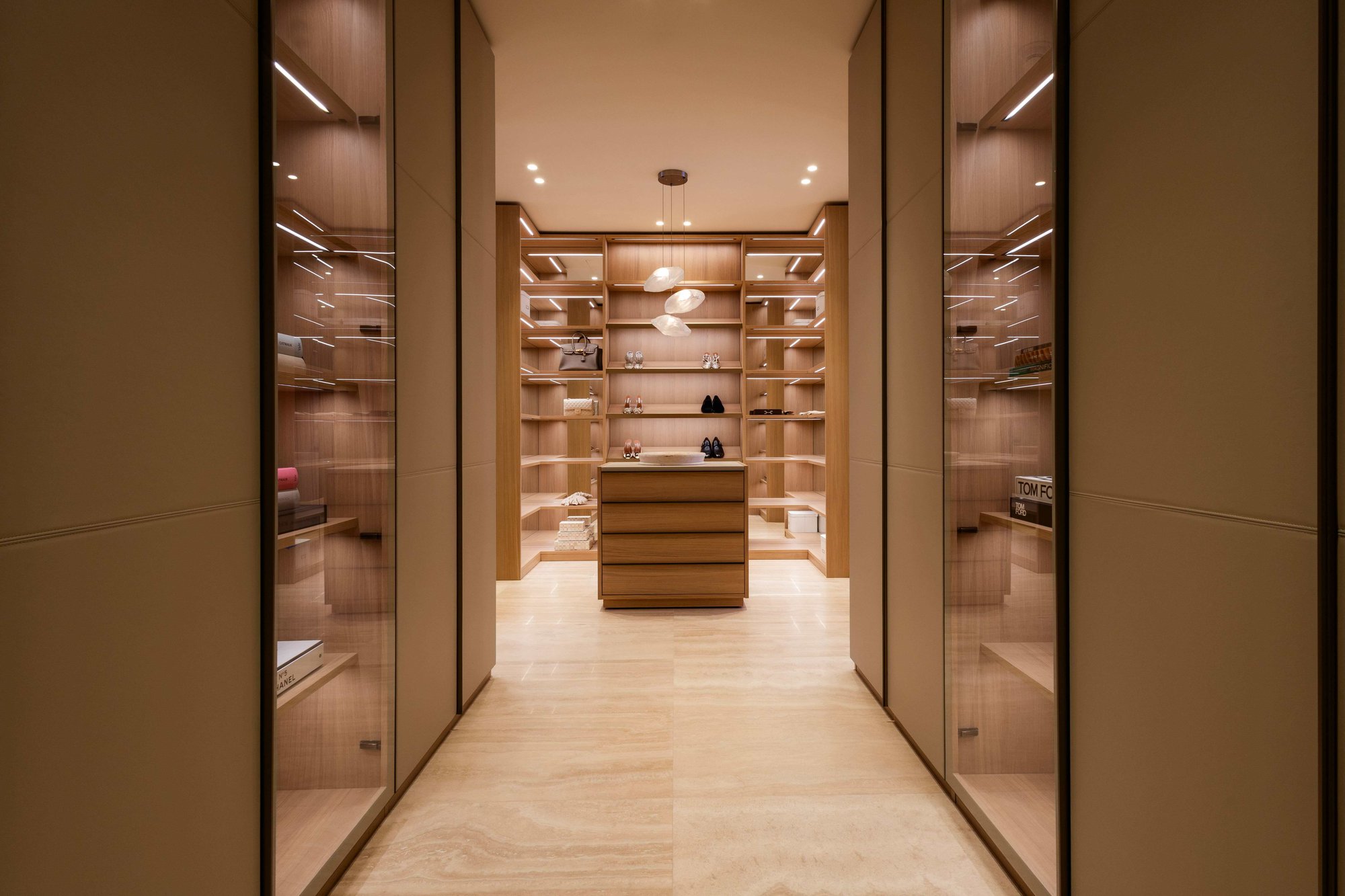 Luxury walk-in closet featuring floor-to-ceiling custom natural wood cabinetry with integrated shelf lighting, and a central dresser island under an organic pendant light. Luxury walk-in closet featuring floor-to-ceiling custom natural wood cabinetry with integrated shelf lighting, and a central dresser island under an organic pendant light.